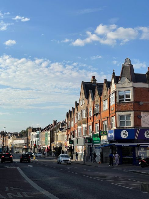 A daytime street scene on Streatham High Road featuring a row of red brick commercial buildings with large windows and signage, including a cafe named 'Carolina' and other local shops. The road has ma